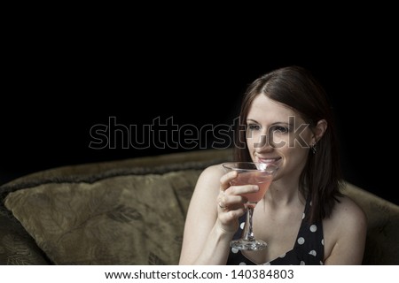 Beautiful young woman with brown hair and eyes in polka dot dress on black background. She is drinking a pink martini.