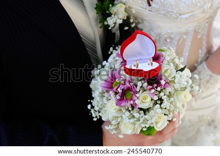Wedding Rings Box; a red wedding rings box on a wedding bouquet with a bride and a bridegroom in bokeh background