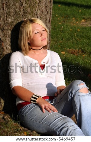 Serious young woman sitting on the ground in front of a tree, her face turned upward toward the sun, and her eyes closed.