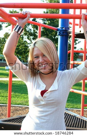 Smiling young blond woman outdoors on a school playground, her arms above her head holding onto climbing bars.