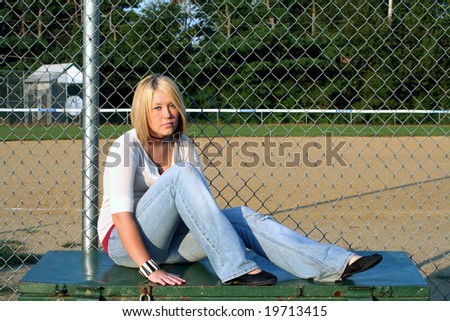 Young blond woman sitting on an outdoor storage container, in front of a chain link fence and baseball field.
