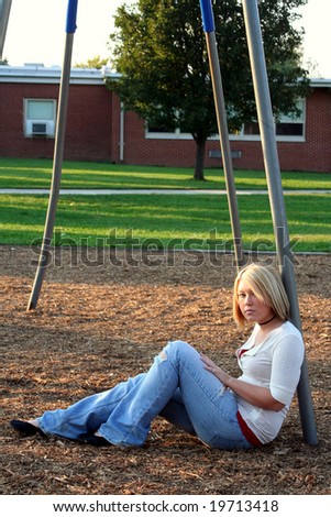 Serious young blond woman sitting on the ground in profile, her face turned to her left toward the viewer, while leaning against a metal pole of a school playground swing set.