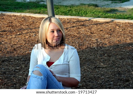 Smiling young blond woman sitting on the ground, her face turned to her left, leaning back against a metal pole of a playground swing set.