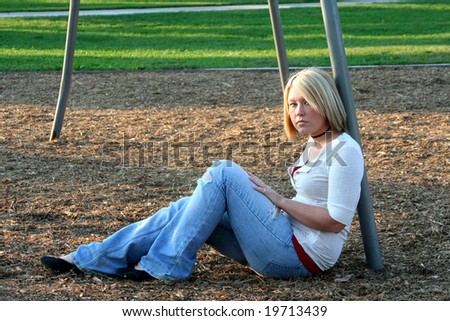 Serious young blond woman sitting on the ground in profile, her face turned to her left toward the viewer, while leaning back against a metal pole of a playground swing set.