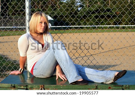 Serious young blond woman sitting on an equipment storage box in front of a chain link fence and baseball field.