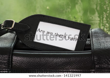 A luggage tag on a black leather suitcase with the phrase Are We There Yet? on the label and a rain soaked window in the background