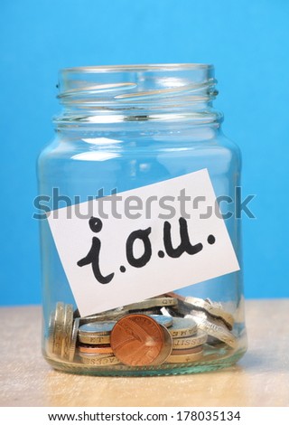 A jam jar part filled with british coins for savings has been slapped with a white sticker that says I.O.U. the abbreviation for I Owe You