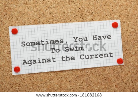 The phrase Sometimes You Have to Swim Against the Current typed on a piece of graph paper pinned to a cork notice board. A concept for having a different vision or point of view from your peers.