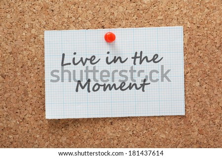 The phrase Live in the Moment printed on a piece of graph paper and pinned to a cork notice board