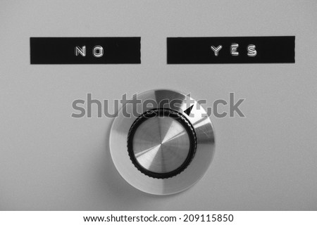 Black and white image of control switch or knob on a metal panel pointing at the printed label for the word Yes