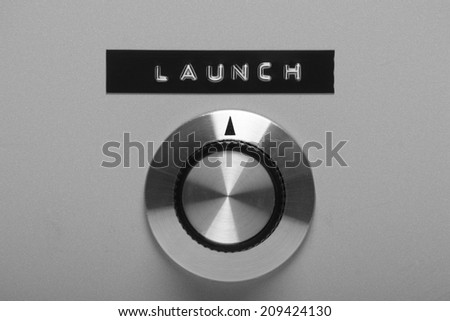Black and white image of a retro style control switch on a metal panel, pointing at a printed label with the word Launch