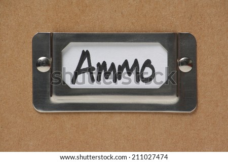 Metal drawer label holder on a cardboard storage box containing a white card with the word Ammo, short for ammunition
