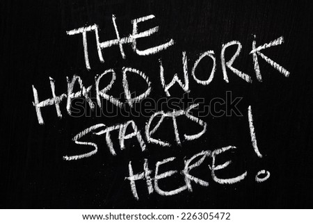 The phrase The Hard Work Starts Here written by hand in white chalk on a used blackboard