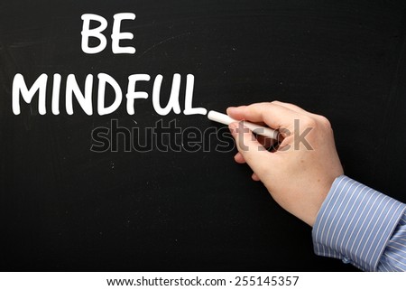 Male hand wearing a business shirt writing the phrase Be Mindful on a blackboard. Being Mindful is a mind and body approach to achieving well-being and reducing stress levels