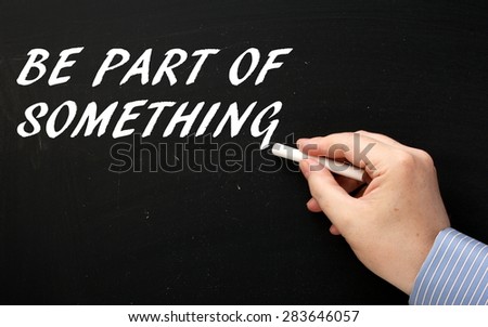 Male hand wearing a business shirt writing the phrase Be Part of Something in white text on a blackboard