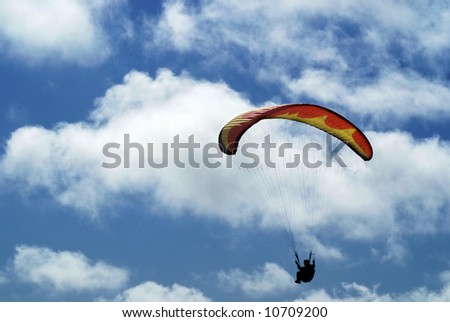 A paraglider with a yellow and orange parachute canopy high in a blue sky with big fluffy white clouds.
