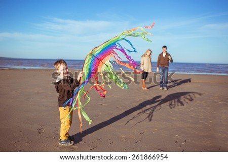 A small boy is holding a rainbow-coloured kite in his hand and his parents are behind him at the background. Clothes: casual.