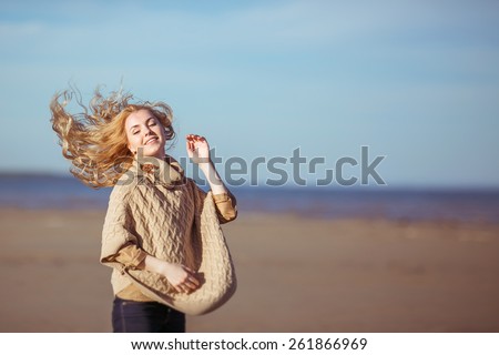A portrait of a young fair-haired woman smiling with the hair waving in the wind. Clothes: a beige knitted poncho, dark-blue jeans.