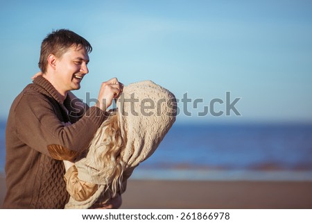 A young dark-haired man is smiling, pulling his wife's knitting poncho on her head, the are at the seashore. Clothes: casual.
