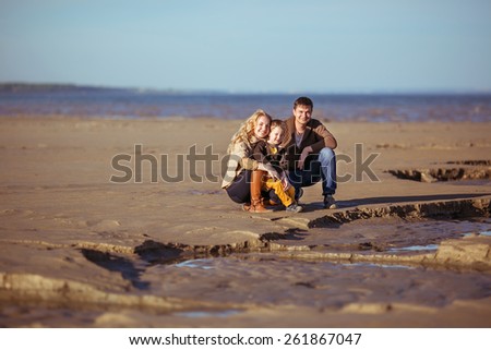 A young family is sitting on the sand in a sunny autumn day and smiling. Clothes: casual