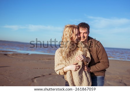 A young man and a young woman are standing close to each other at the beach and smiling. Clothes: casual.