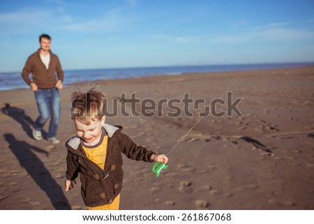 A small boy and his dad are running on the sand in a sunny day and flying a kite. Clothes: casual.