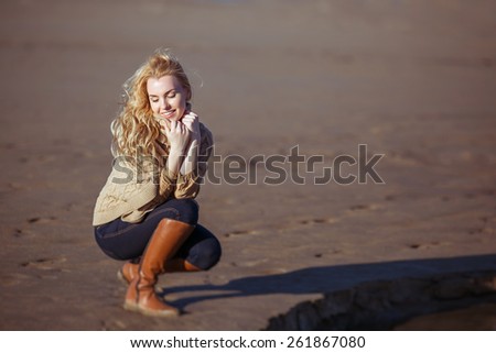 A portrait of a young fair-haired woman sitting on her haunches on the sand and smiling with her eyes closed. Clothes: a beige knitted poncho, dark-blue jeans, brown boots.