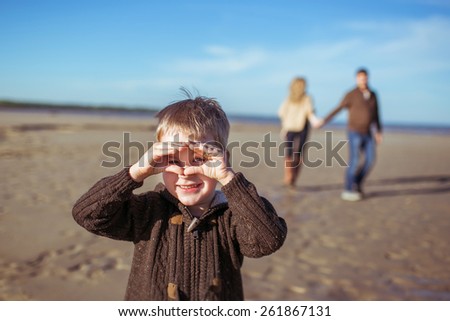A portrait of a five-year-old boy holding hands like a mask at the beach on a sunny morning and his  parents coming hand-in-hand.
