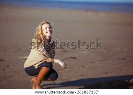 A young fair-haired woman is sitting on her haunches on the sand in a sunny day and smiling. Clothes: a beige knitted poncho, dark-blue jeans, brown boots.