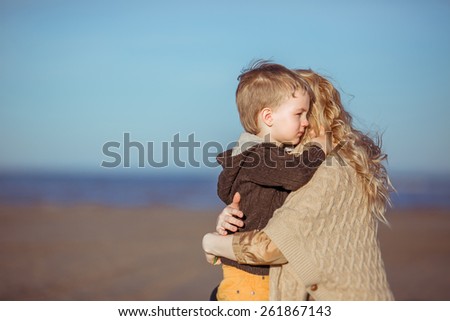 A five-year-old boy and his mother are embracing at the seashore in a sunny day. Clothes: casual.