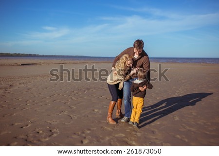 Smiling parents and their five-year-old son are embracing one another at the beach. Clothes: casual.