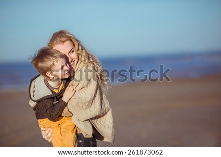 A young fair-haired woman is keeping her five-year-old son on her hands and smiling. Clothes: casual.