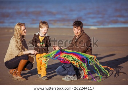 The family is going to fly a rainbow-coloured kite at the seashore. Clothes: casual.