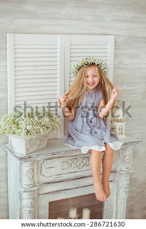 A young girl is sitting on the fireplace and tiding her hair. She is grinning all over her face. She is having a floral wreath and casual clothes on. The atmosphere of happiness is all around her.