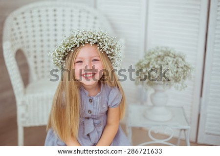 A young girl is grinning all over her face. She is wearing a light blue dress and a floral wreathe on. The armchair and flowers are behind the girl.