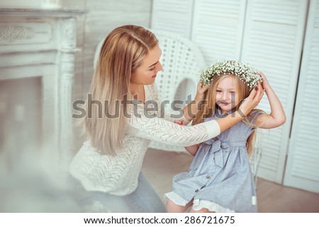 ? happy mother is taking care of her young daughter. She is trying to put a floral wreath on her. A mother is having jeans and white blouse on.The atmosphere of happiness is all around them.