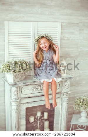 A young girl is sitting on the fireplace and tiding her hair. She is grinning all over her face. She is having a floral wreath and casual clothes on. The atmosphere of happiness is all around her.