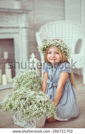 A young warm smiling girl is sitting on the wooden floor near the flowers. She has a light blue dress and a floral wreath on. The atmosphere of happiness is all around her.