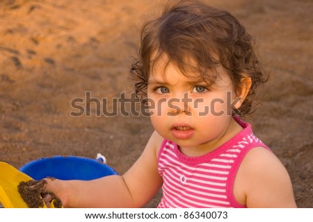 Horizontal upper body shot of a beautiful Caucasian toddler girl playing in sand with a yellow scoop and a blue bucket.