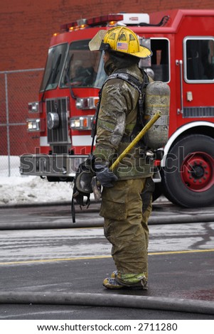 A winter scene of a fireman watching a fire with a fire truck in the background.