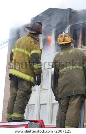 Two firemen standing on a fire truck watching an apartment fire.
