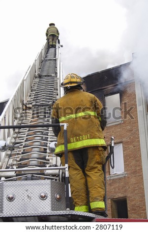 A Fireman standing at the bottom of the ladder and a fireman on the top fighting the fire.