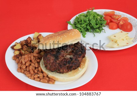 Thick hunk of hamburger mixed with pieces of jalapeno pepper and other spices, char-grilled and served with baked beans, potatoes and selected fixings against bright red background.