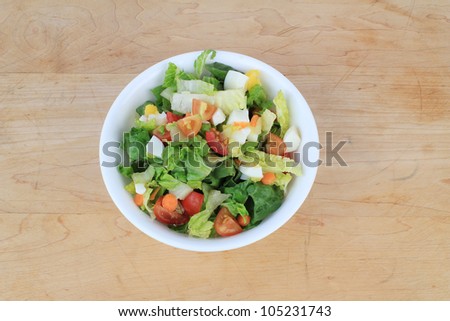 Salad ingredients in white bowl on old wooden cutting board.  Includes boiled eggs, tomatoes, carrots, lettuce, onions, raisins.