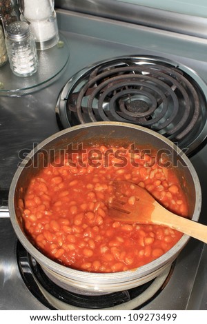 Spicy BBQ beans in saucepan with wooden spoon cooking on electric stove.