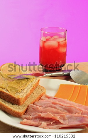 Putting together a ham and cheese sandwich on honey wheat bread with brown mustard.  Glass of ice cold fruit punch, knife and cheese slicer.