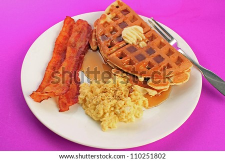 Breakfast plate with buttered waffle and syrup; fried bacon; and scrambled eggs on purple background.