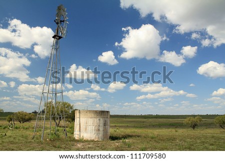 Windmill and water tank on Central Texas Ranch with brilliant blue sky and puffy clouds and typical vegetation of scrub brush and cactus.