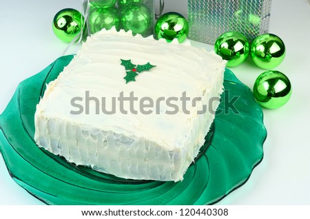 White Christmas Cake with holly leaf decoration on top sitting on green platter and surrounded by green Christmas Tree Bulbs and gifts.