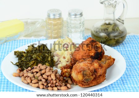 Black eyed peas and turnip greens with deep fried chicken legs and potatoes and spicy vinegar sauce on blue gingham place mat.  Southern cooking where dinner is often called supper.